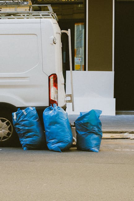 A black wheeled waste bin with a white label reading 'St. John's' sits on a paved sidewalk at night, partially filled with discarded cardboard boxes and various rubbish items. The lid is open, reveali